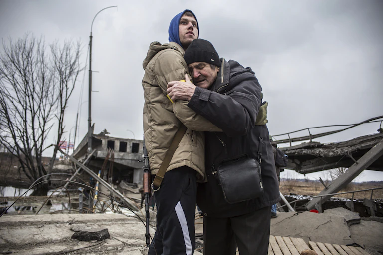 Two men embrace near a ruined bridge.