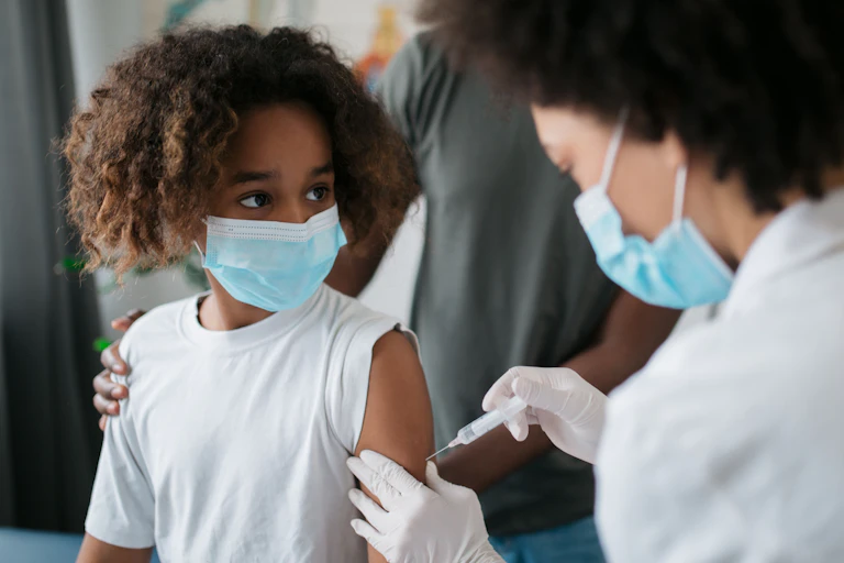 A girl getting vaccinated by a female doctor, both of whom are wearing face masks.