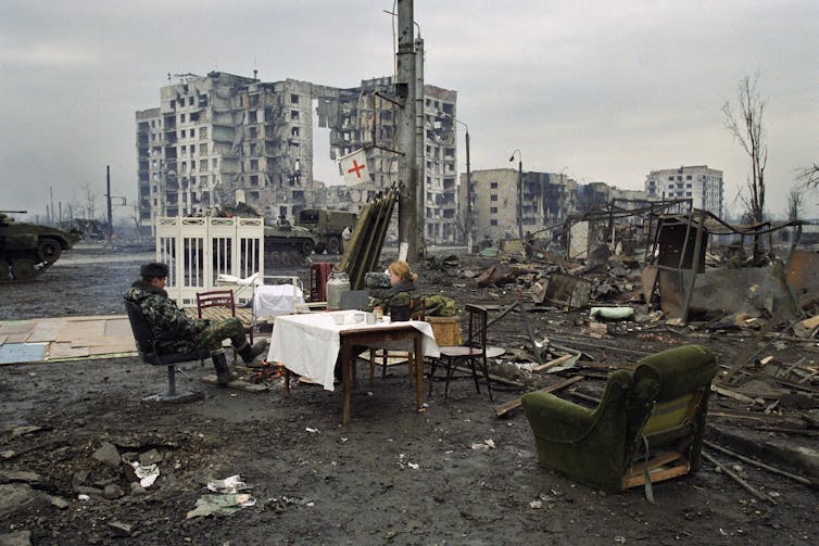 Russian soldiers sit at a table with a white table cloth amid bombed ruins.