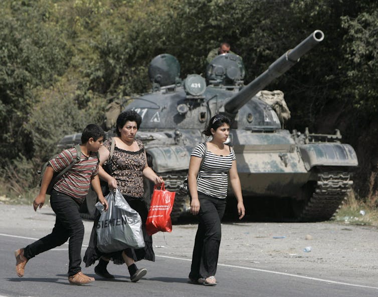 A dark-haired woman and two children walk past a tank