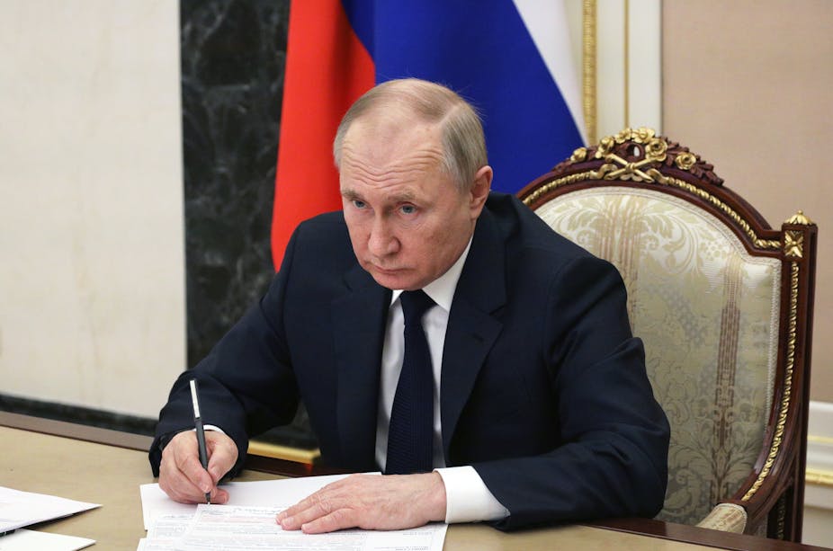 Russian president Vladimir Putin sits at a table in the Kremlin writing notes and looking at a video