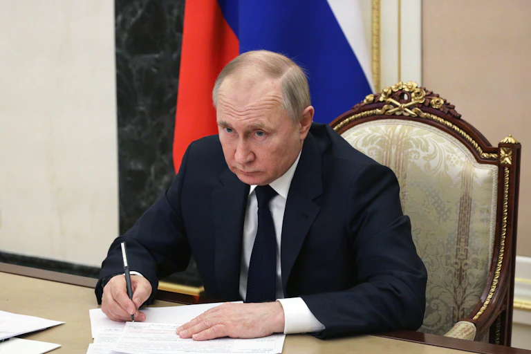 Russian president Vladimir Putin sits at a table in the Kremlin writing notes and looking at a video