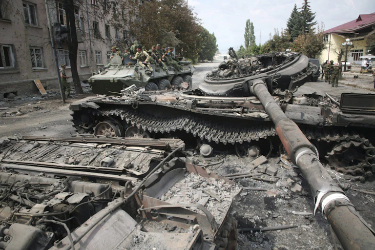soldiers ride on top of a tank with a destroyed tank in the foreground