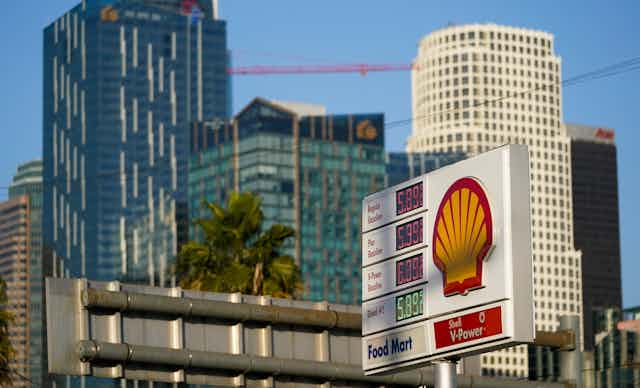 A gas station sign with a shell logo on it displays the prices of gas above 5 dollars in front of several tall buildings