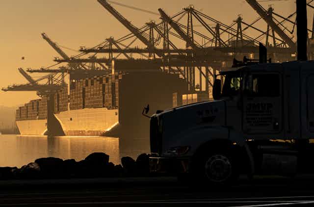 The cab of a truck is in the foreground as two packed cargo ships sit in the water at a port, with cranes hanging above them