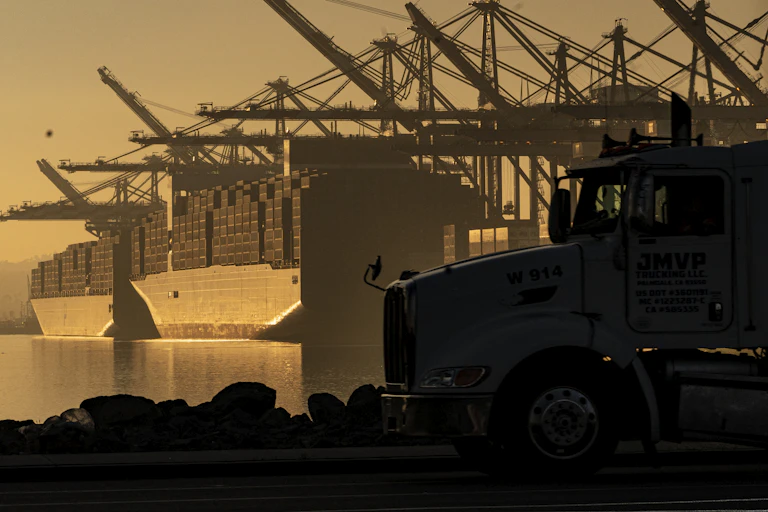 The cab of a truck is in the foreground as two packed cargo ships sit in the water at a port, with cranes hanging above them