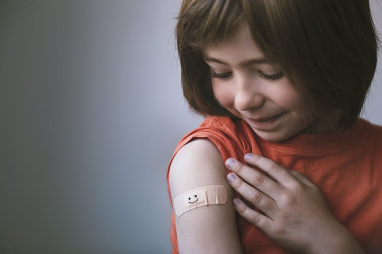 A girl with bandaid on her bicep at the doctor's office. A smiley face is drawn on the bandaid.