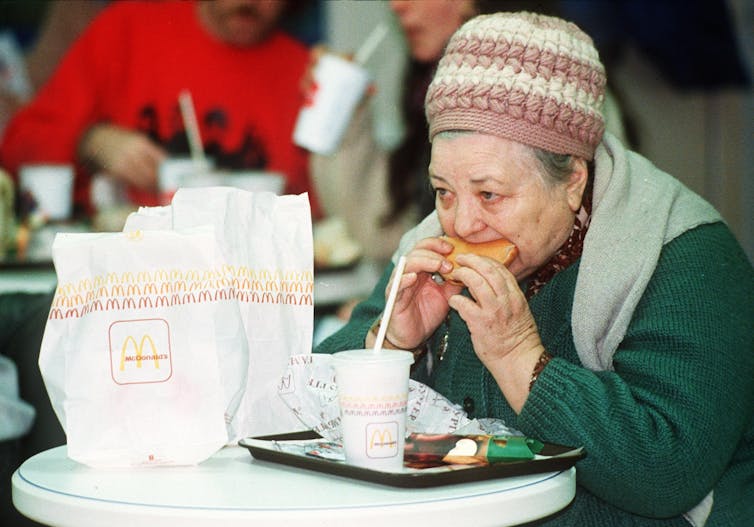 An elderly white Russian woman with a pink and white hat eats a hamburger with McDonalds bags and a drink on a table in front of her