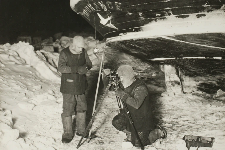 A black-and-white photo of two men in winter clothing standing on ice with a wooden ship behind them