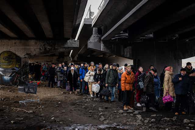 A darkly lit photo shows a crowd of people in coats standing in rubble, under a split bridge 