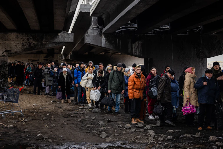 A darkly lit photo shows a crowd of people in coats standing in rubble, under a split bridge