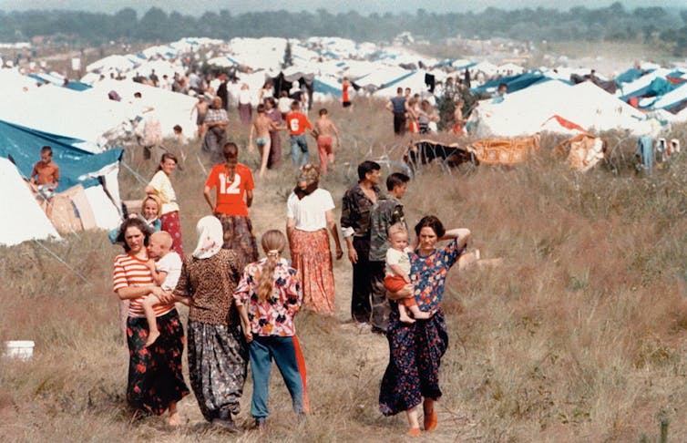 A crowd of women holding children and young people are seen in front of rows of tents