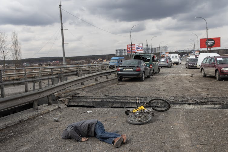 The body of a person lies on the ground of a destroyed bridge, with cars parked in the foreground.