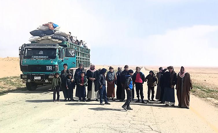 A row of people stand next to an open truck, filled with bags and people. An empty dirt road is behind them.