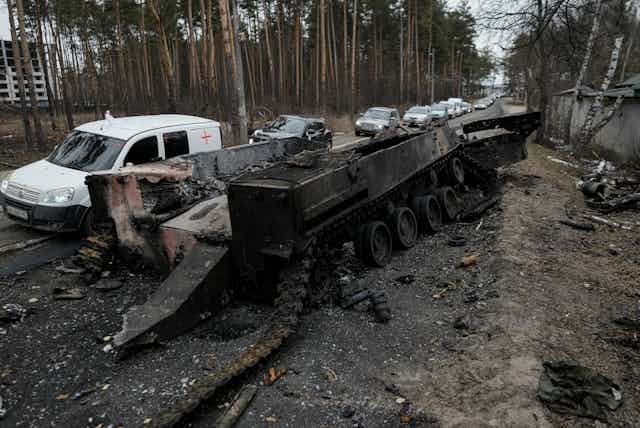 Vehicles cross a road, passing a destroyed Russian tank on the side. 
