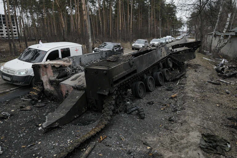Vehicles cross a road, passing a destroyed Russian tank on the side.