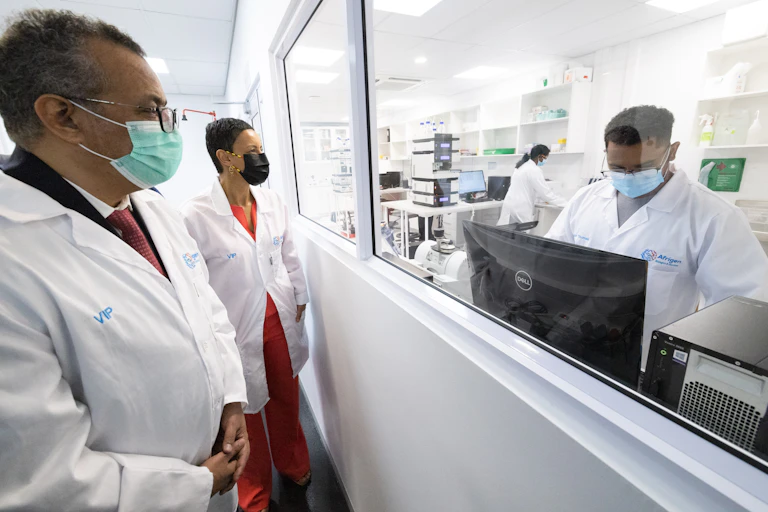 Man looking at another through a window. They're both wearing white lab coats and face masks.