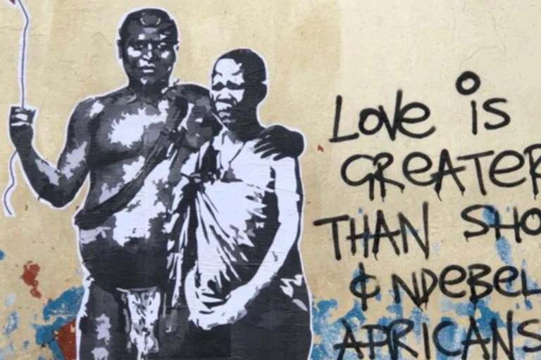 A street art murall showing a man in traditional attire with his arm around a woman, also in traditional attire. He holds a string with a red balloon at the end of it. Next to them the words 'Love is greater than Shona & Ndebele Africans.