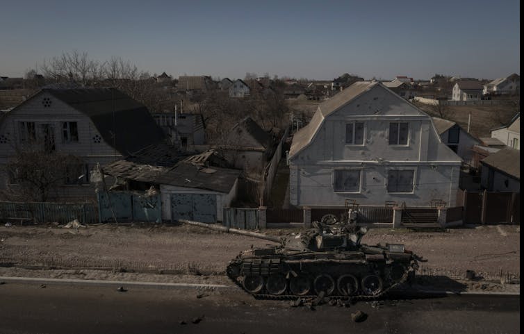 A destroyed tank sits in front of damaged and destroyed buildings.