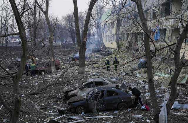 Emergency workers walk among the smoky ruins of a building and burnt cars and trees