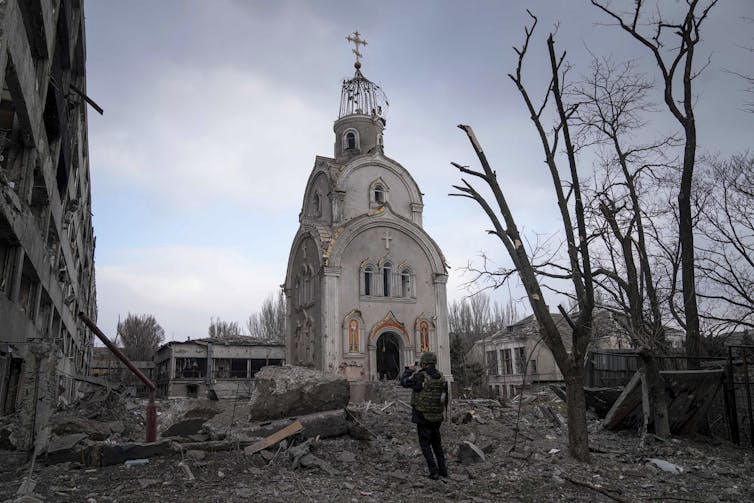 a soldier stands in rubble and takes a photograph of a damaged church