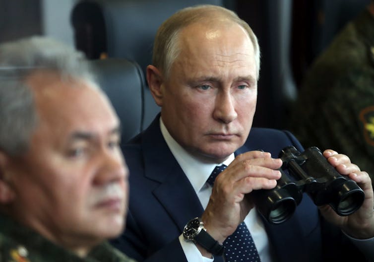 A bald man holds binoculars while another man with grey hair sits next to him.