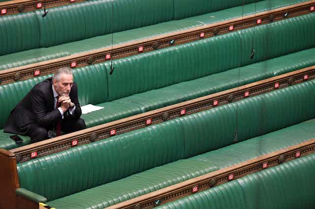 A member of the British Parliament sitting alone on the green benches of the House of Commons during social distancing in the pandemic. 