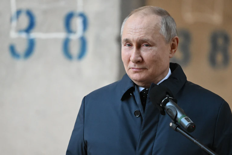 A balding man in a dark trenchcoat standing in front of a microphone.