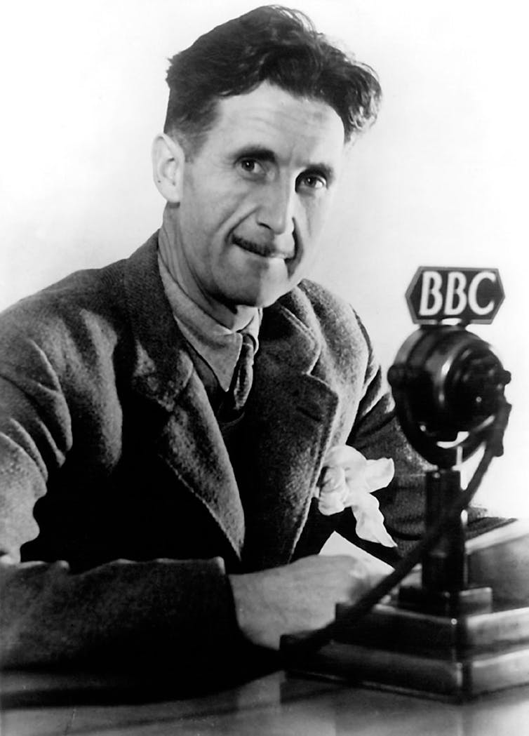 A middle-aged man with dark hair and wearing a tweed jacket, vest and tie, sits before a microphone that says BBC on it.