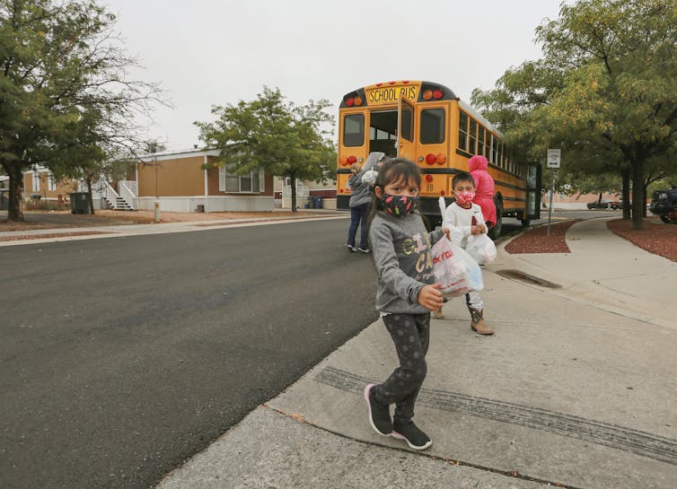 Children near a school bus, wearing masks, carry bags of food.