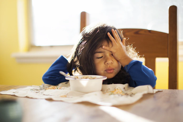 sleepy-looking toddler at breakfast table rubs face