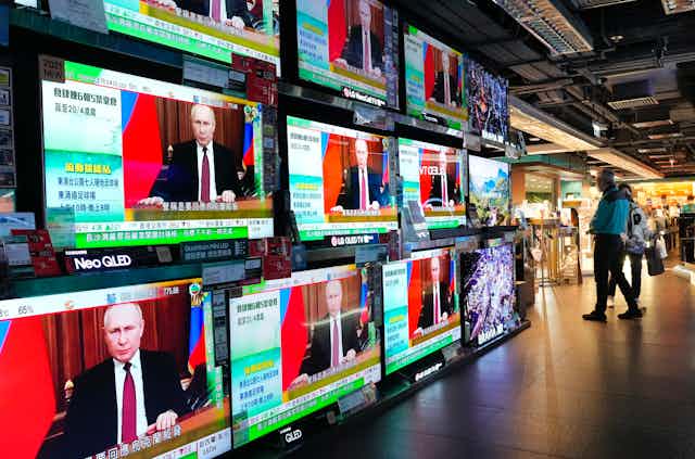 A wall of television screens display a man in a dark business suit with a red and blue flag behind him