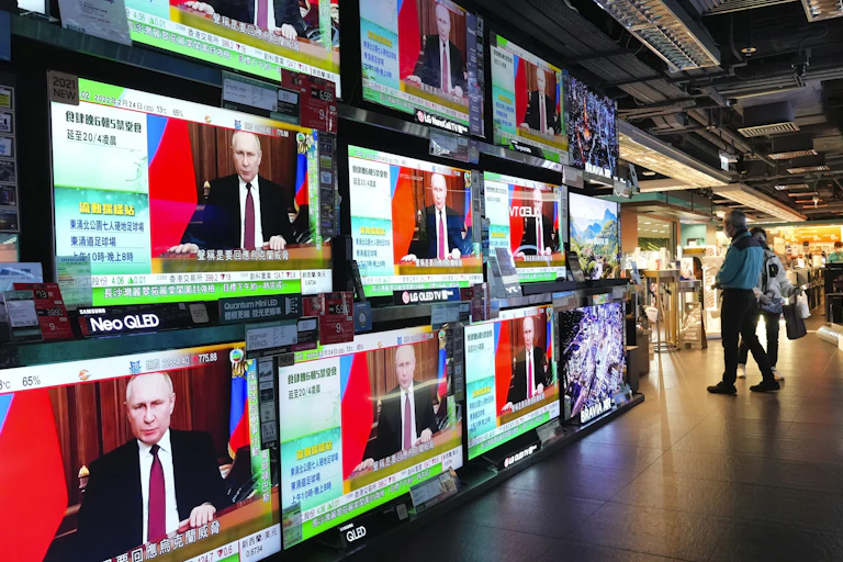 A wall of television screens display a man in a dark business suit with a red and blue flag behind him