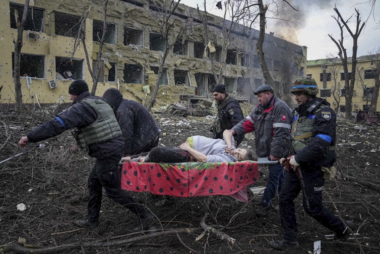five men carry a pregnant woman on a stretcher across a plaza strewn with tree limbs with smoke and bomb blasted buildings in the background