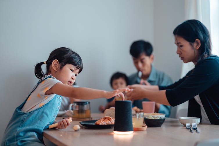 a toddler touches the top of a black cylinder on a dining table as a family eats in the background