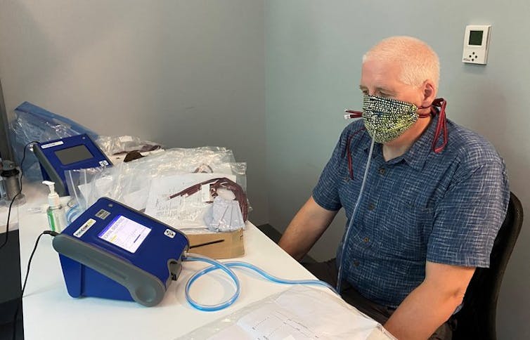 A man wearing a cloth face mask with a tube extending from it to a machine on the table in front of him