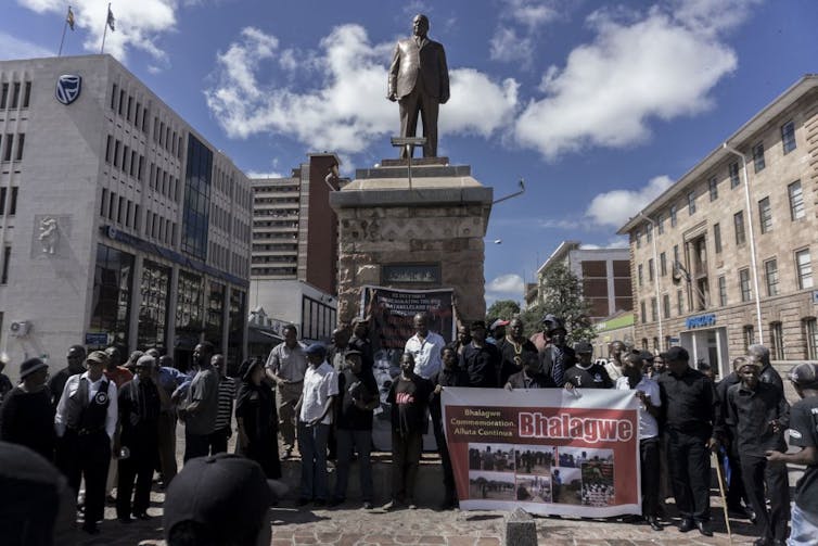 A crowd with banners gathers around a statue of a man standing proudly on a plinth.