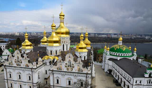Towers and gold domes with crosses seen atop a white monestary building complex in front of water.