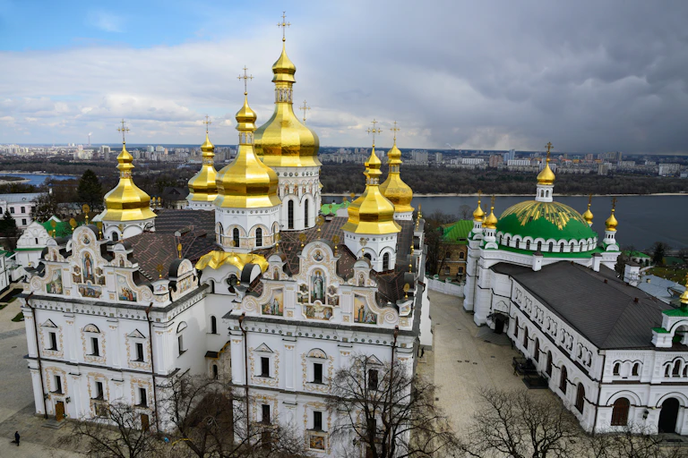Towers and gold domes with crosses seen atop a white monestary building complex in front of water.