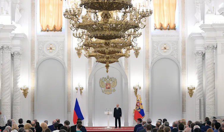 A man stands under an ornate golden chandelier in a palace with white walls and golden embellishments..