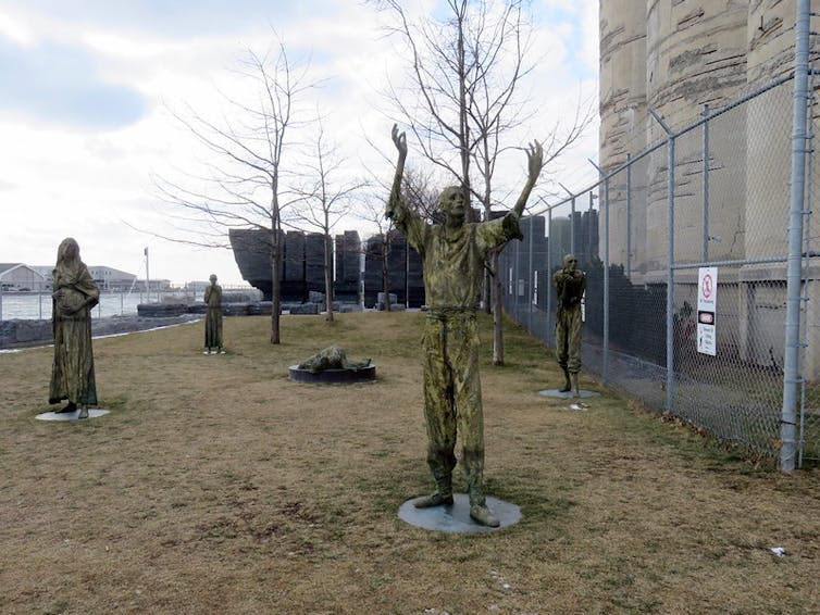 Statues of emaciated figures seen in front of large slabs of stone in the background.