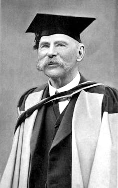A man in an academic gown and bushy moustache. 