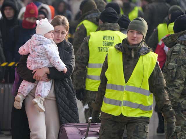 A woman with dark blond hair carrying a baby girl walks beside a Polish aid worker who pulls her luggage.