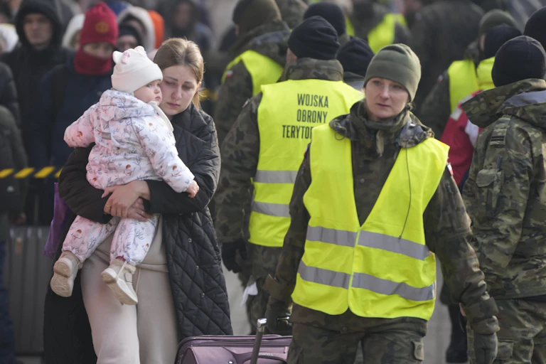 A woman with dark blond hair carrying a baby girl walks beside a Polish aid worker who pulls her luggage.