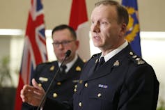 Two men in uniforms stand with a podium and a microphone in front of them and flags behind them.