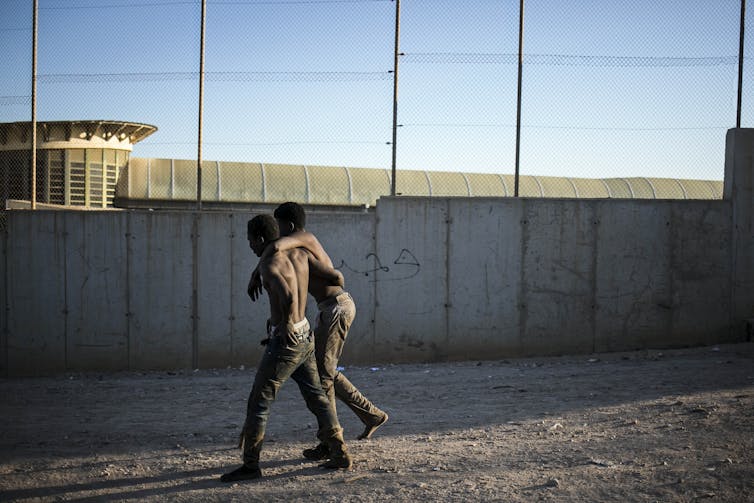 One Black man leans on another with a concrete fence in front of them.