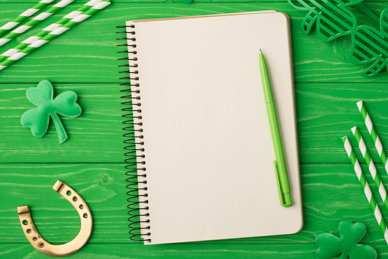 A blank notebook and green pen seen against a green table and shamrocks.