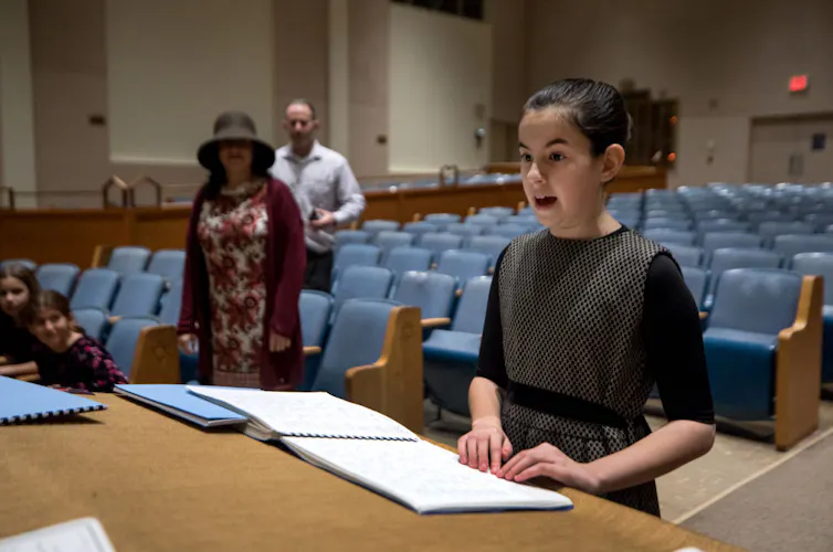 A girl chants from a religious text inside a synagogue as two adults look on.
