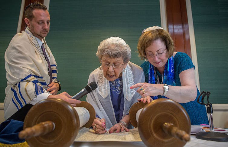 A man and woman stand on either side of an elderly woman reading from a Torah scroll.