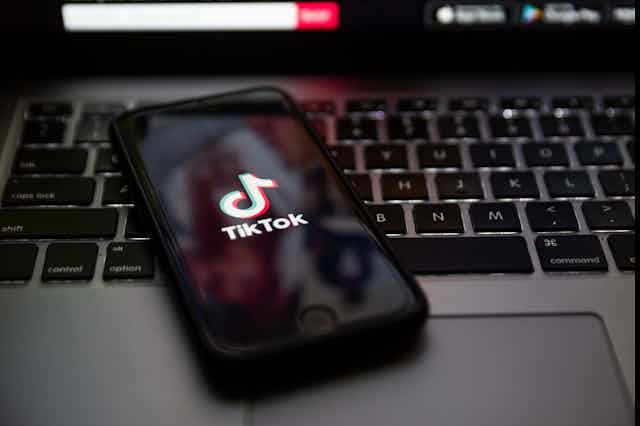 a phone showing the tiktok logo rests on an apple laptop keyboard
